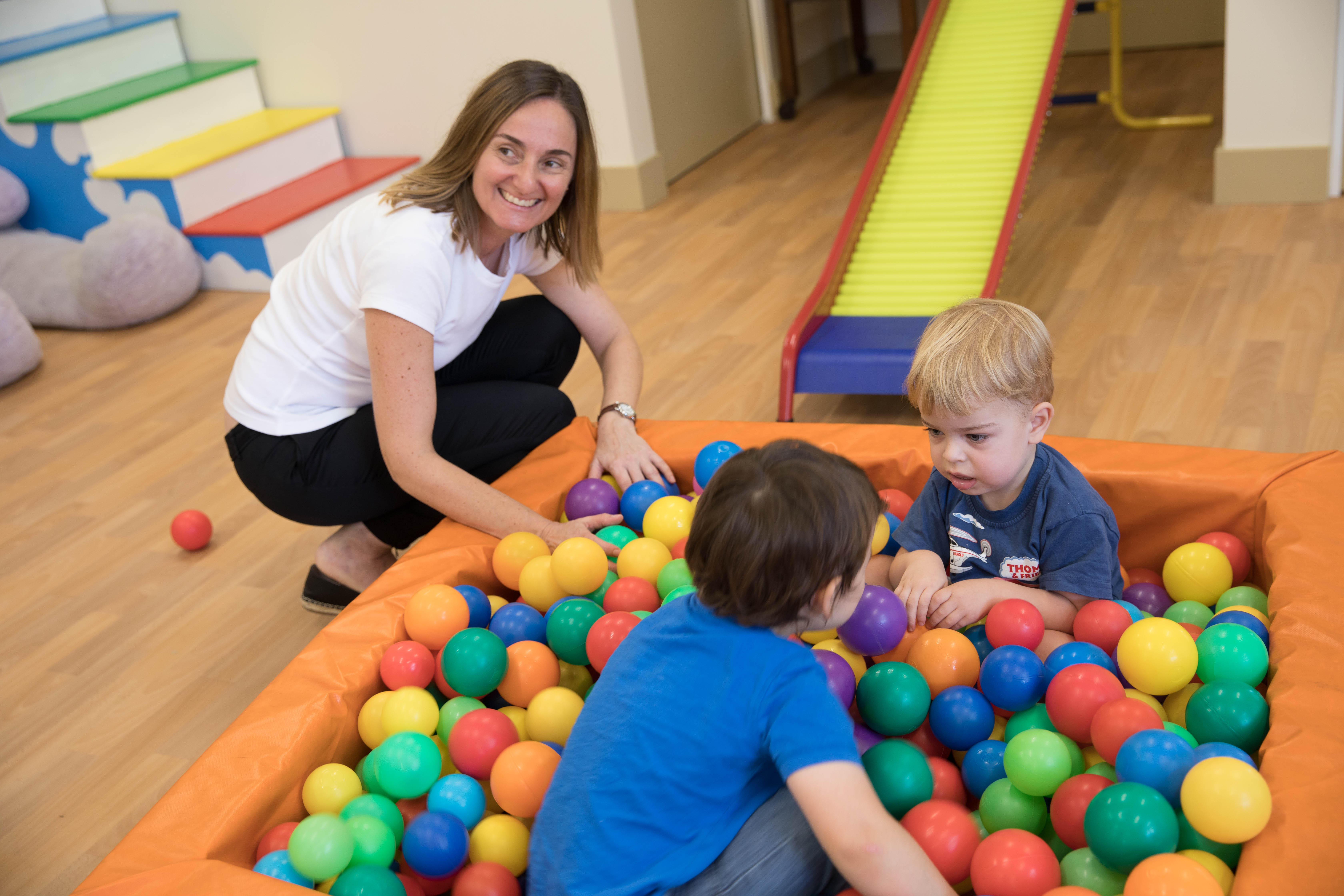 A woman crouches down next to a playpen full of plastic, coloured balls. Two young boys play in the playpen. 