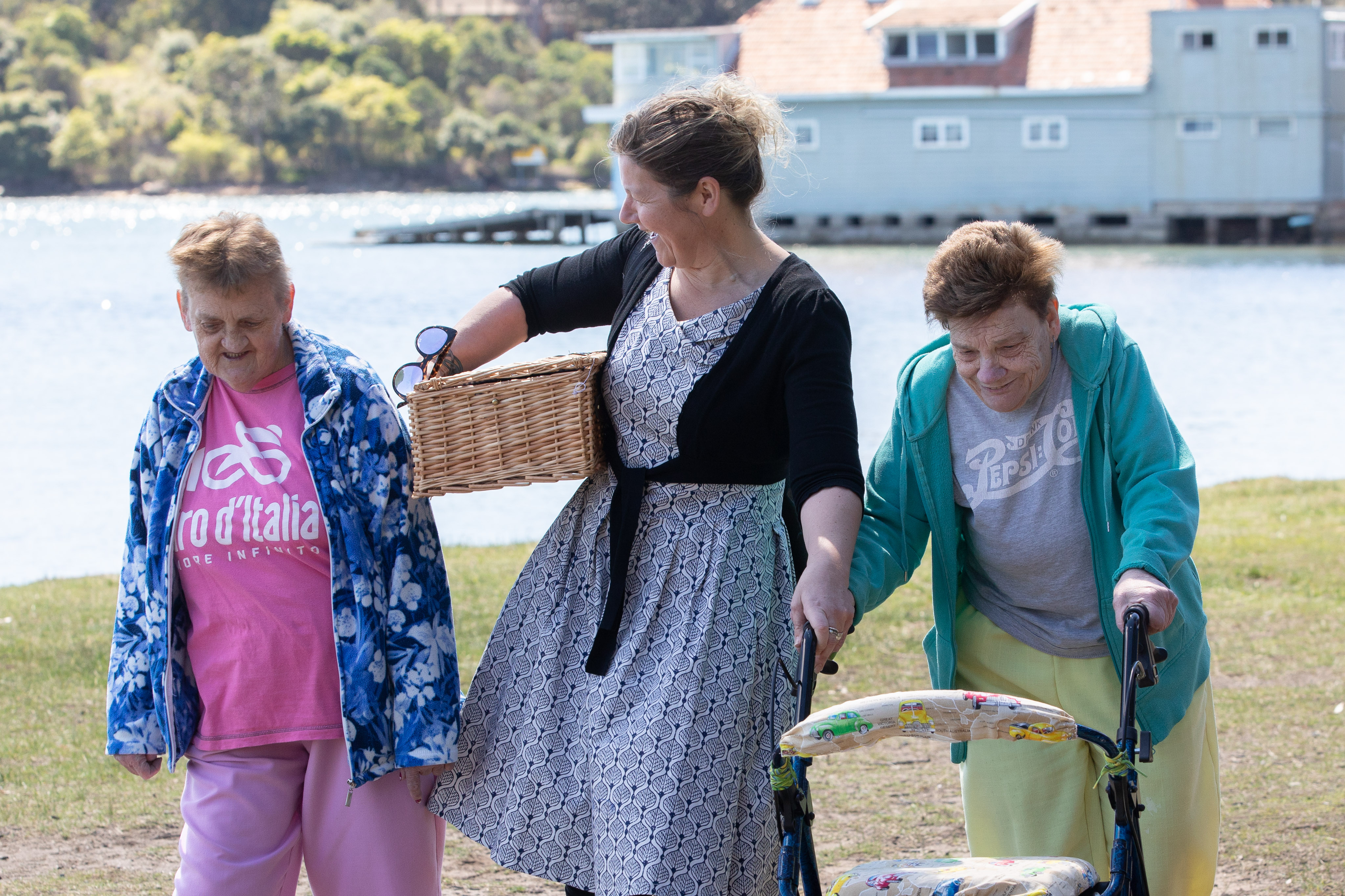 Three women are walking on a sunny day, chatting. They are walking past a bay, one woman is carrying a picnic basket, another is using a walker to assist her. 