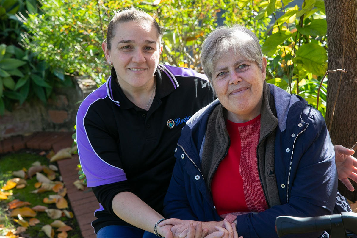 Two women sit outside in a lush, green surrounding. One is wearing a purple shirt, with her hair tied back. The other woman wears a dark jacket and has short, grey hair. Both women smile and look at the camera.