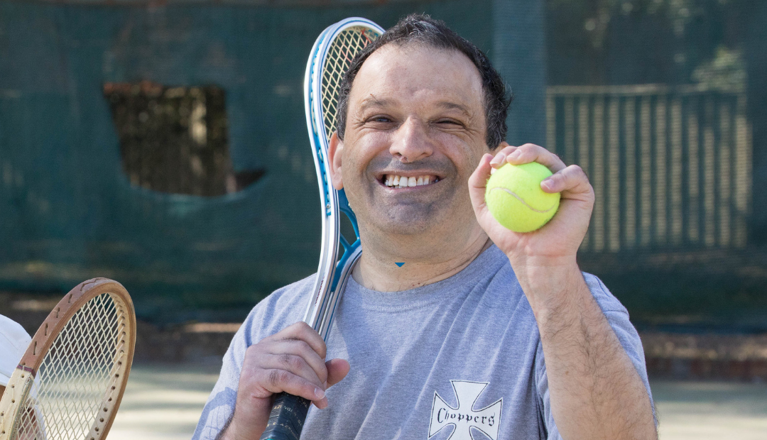 A man smiling. He has a tennis racket over one shoulder and a tennis ball in the other.